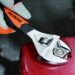 Gloved hand using a WOKIN stubby adjustable wrench to tighten a nut on a red metal surface.