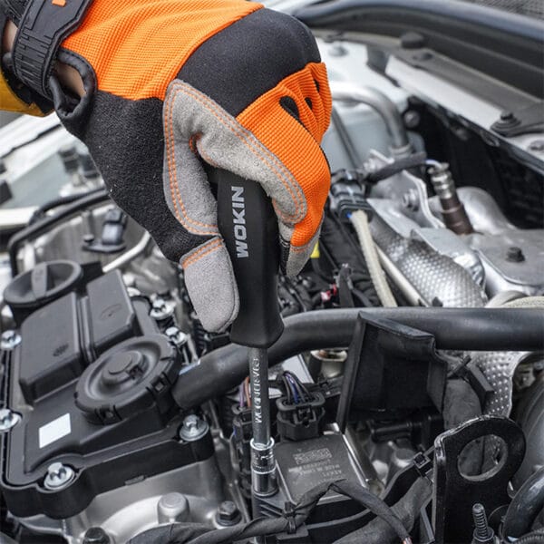 Hand wearing WOKIN work glove using WOKIN screwdriver and socket to work on car engine, close-up